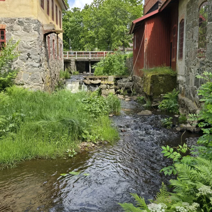 Historisk bruksmiljö på Huseby Bruk med rinnande vatten, äldre industribyggnader och lummig grönska.